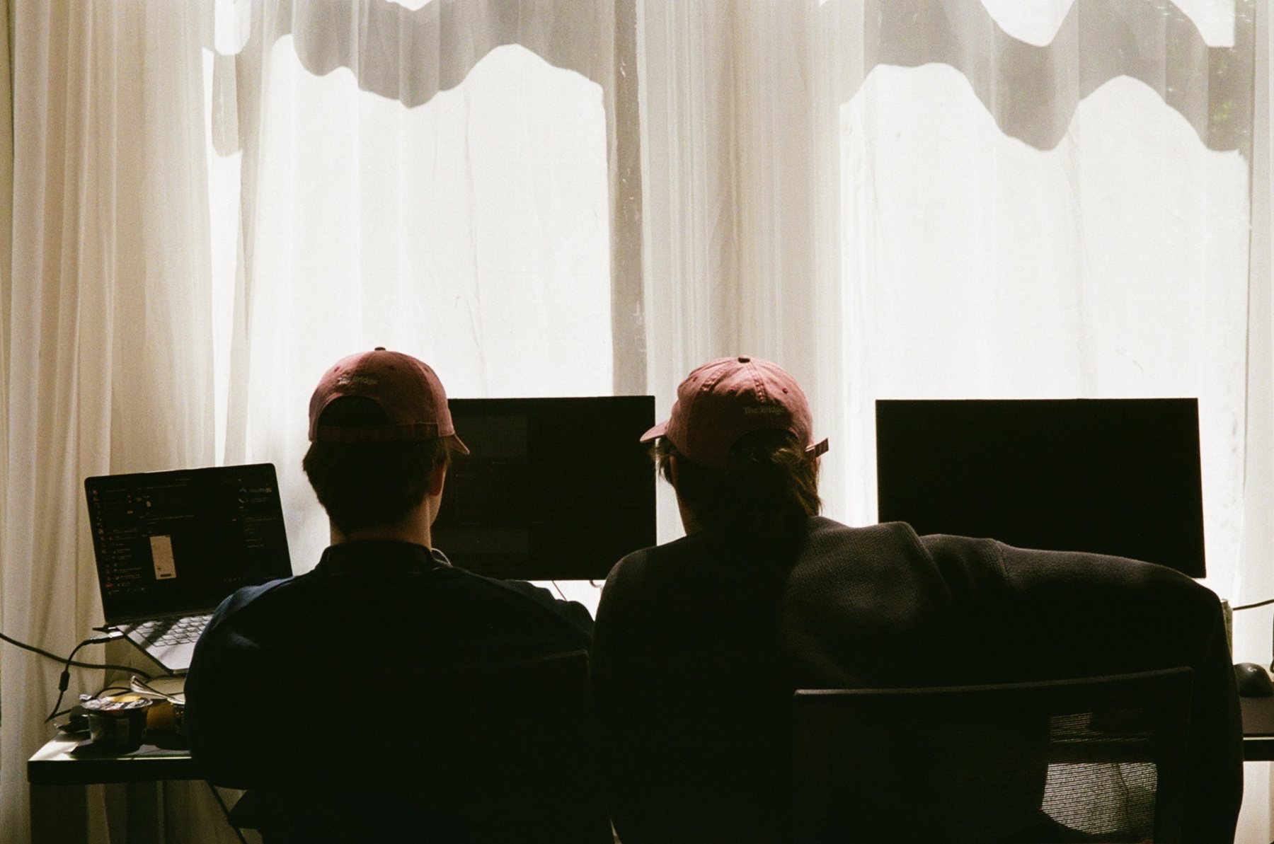 Two founders in The Bridge caps working in front of bright curtains